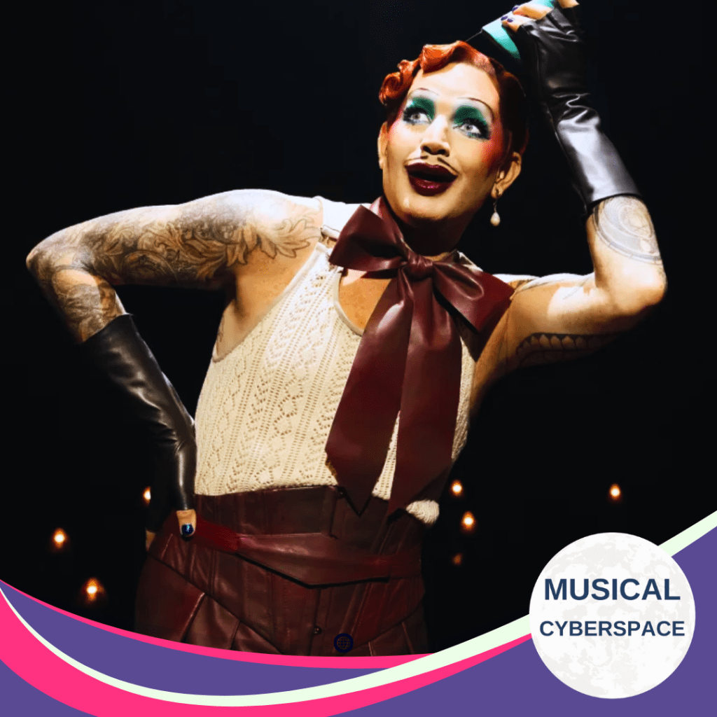 Adam Lambert as the Emcee in Cabaret, striking a theatrical pose in bold makeup and costume under dramatic stage lighting, with Musical Cyberspace branding overlay.