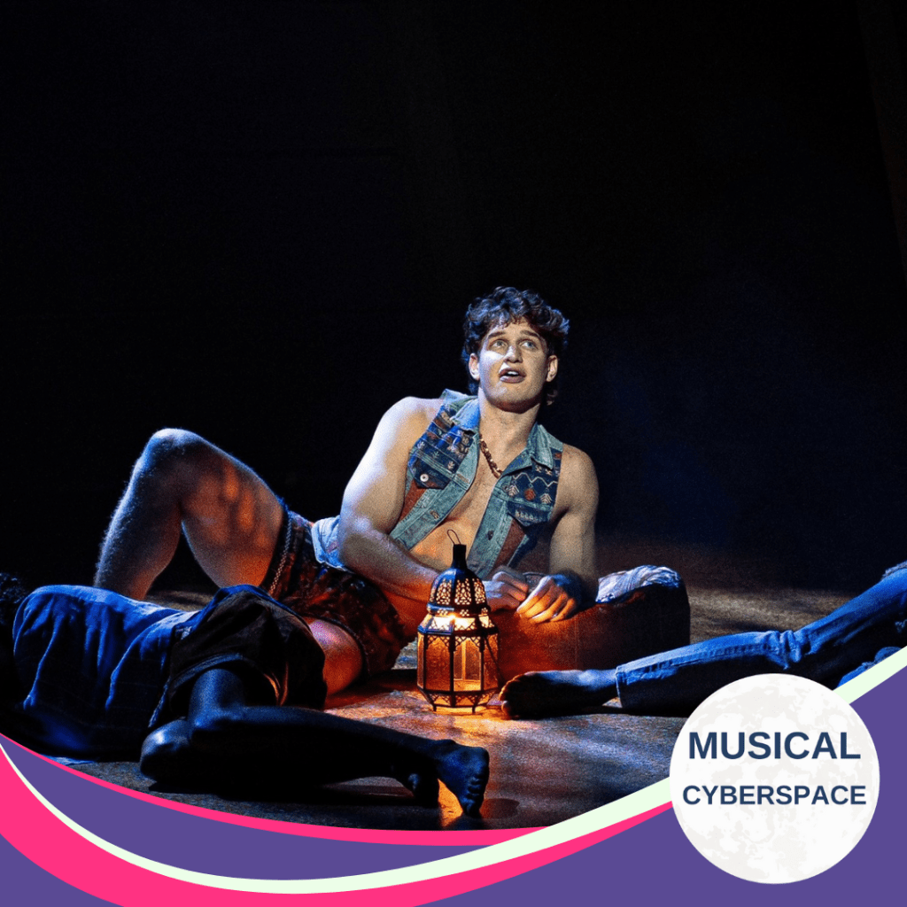 Dylan Janse van Rensburg as Joseph sits onstage under a glowing lantern, wearing a sleeveless denim vest and technicolor shorts, surrounded by castmates in dramatic low lighting.