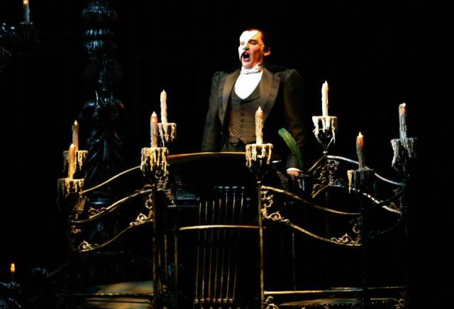 A dramatic scene from a stage performance featuring a male performer in a formal black suit and bow tie, passionately singing from a dark, ornate set with candles, evoking a gothic atmosphere.