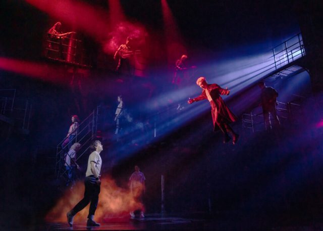 A dramatic theatrical scene featuring actors on stage with colorful lighting and fog effects, showcasing a moment of conflict and tension.