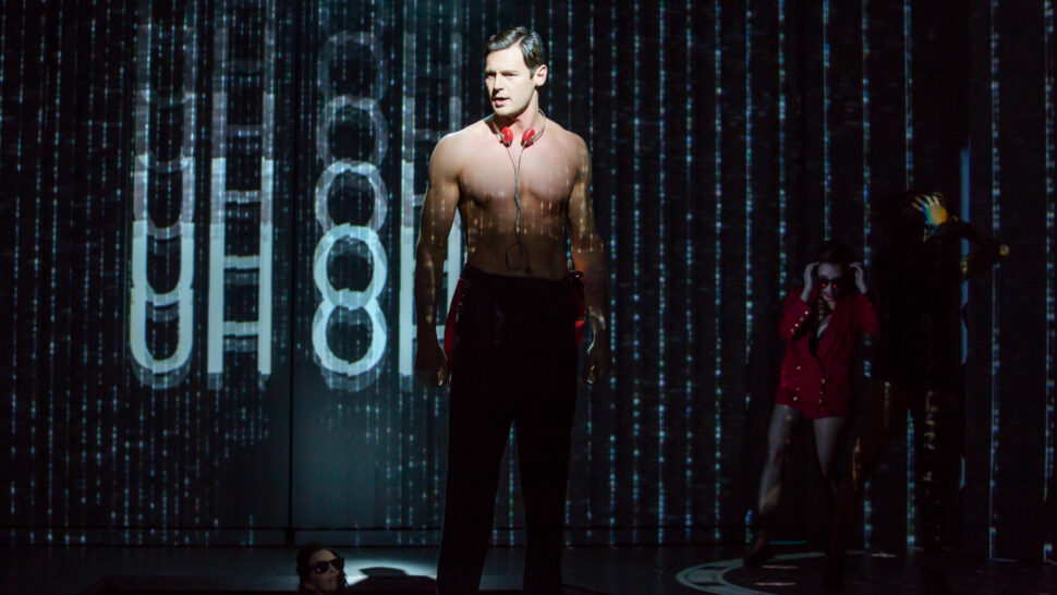 A shirtless male performer stands on stage in a dramatic pose, with digital projections in the background. One performer in a red outfit appears in the shadows, suggesting a theatrical scene.