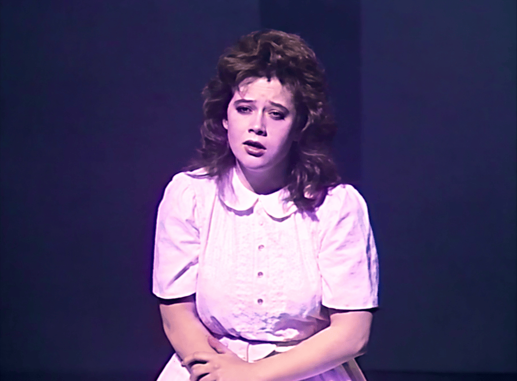 A young woman in a vintage white dress with a collar looks distressed while standing on stage under dim lighting.