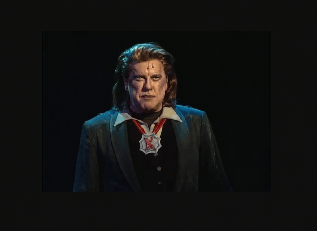 A man in a dramatic costume with long hair and a grim expression, wearing a brooch with a star-shaped design.