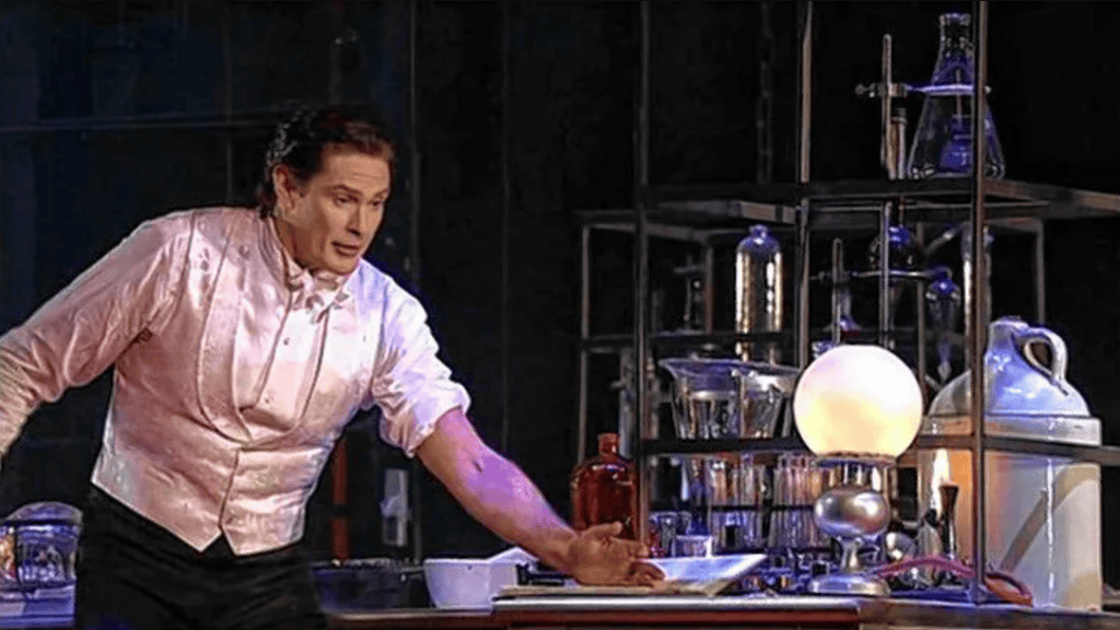 A performer in a pink jacket demonstrates a scientific experiment on stage, surrounded by various laboratory glassware and equipment.