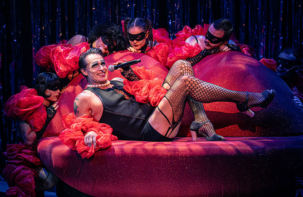 A performer lounging on a large, glittery red cushion, wearing a black outfit and fishnet stockings, holding a microphone while surrounded by several masked dancers in vibrant costumes.