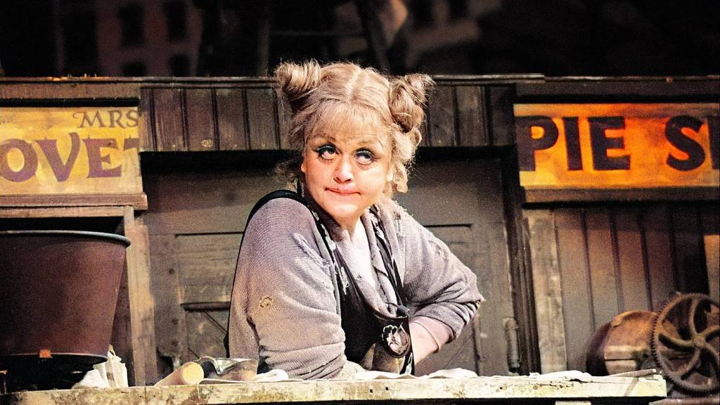 A woman with playful hair styled in buns, wearing a gray outfit and apron, stands behind a wooden counter with signs reading 'Mrs. Lovett' and 'Pie Shop.' She has expressive blue eyes and a quirky smile.